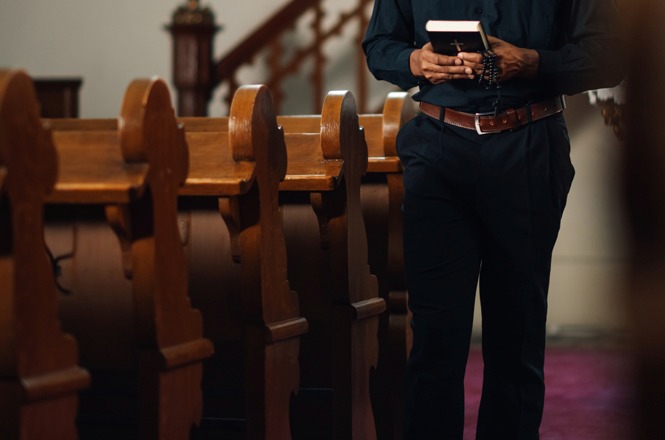 Pessoa segura Bíblia e terço entre os bancos de uma igreja, em referência à prática religiosa.