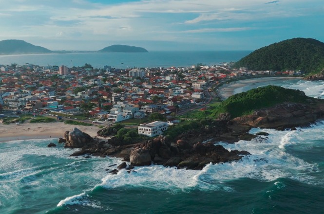 Vista aérea do litoral catarinense mostra área urbana, praias, costões e o mar sob céu parcialmente nublado.