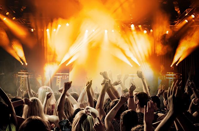 Público acompanha show com mãos erguidas diante de palco iluminado por refletores e efeitos de luz.
