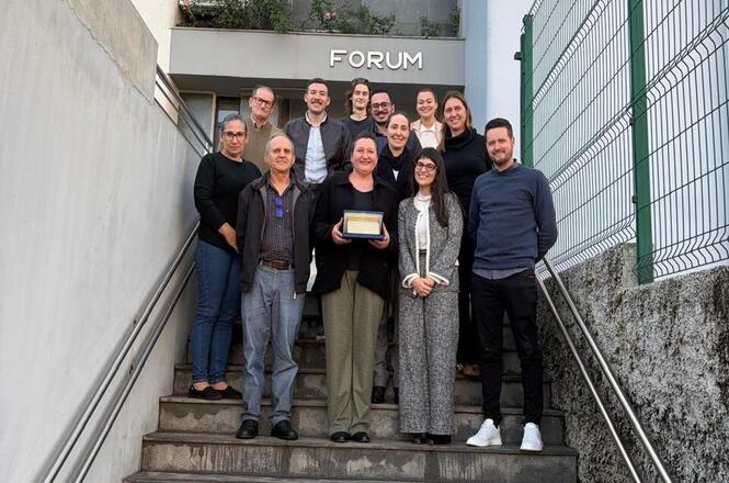 Servidora homenageada pela aposentadoria posa com colegas em frente ao fórum, segurando placa.