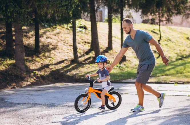 Pessoa adulta acompanha criança aprendendo a andar de bicicleta em área arborizada.