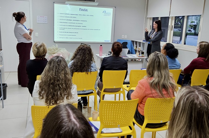 Foto horizontal: Servidoras acompanham apresentação durante reunião de trabalho com exposição de pauta em tela de projeção.