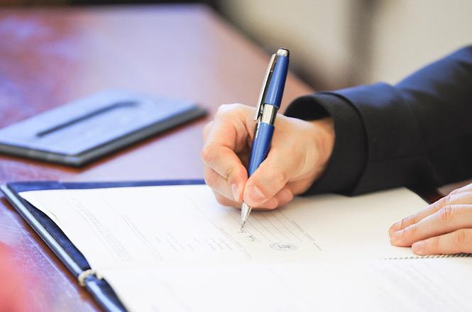 Foto horizontal: detalhe de uma pessoa assinando documento com caneta sobre mesa, em imagem ilustrativa que representa formalização de atos, contratos e procedimentos administrativos.