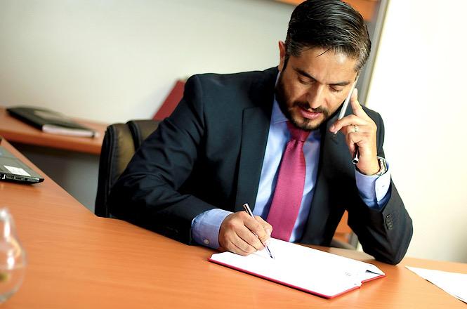 Foto horizontal: homem de terno fala ao telefone enquanto faz anotações em documento sobre a mesa, em imagem ilustrativa que representa atendimento, gestão administrativa e atividades profissionais.
