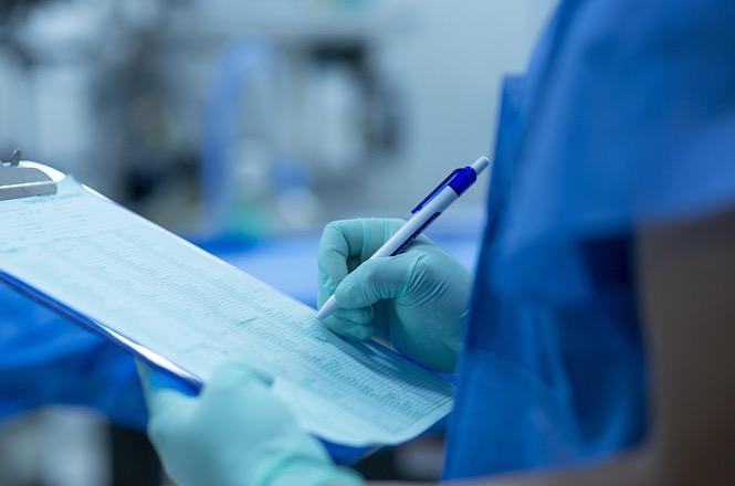 Foto horizontal: profissional de saúde, utilizando luvas, registra informações em prontuário ou ficha clínica durante atendimento