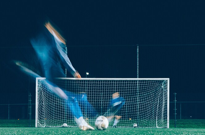 Foto horizontal: jogador chuta uma bola em direção ao gol durante treino noturno de futebol, em imagem com efeito de movimento que destaca a velocidade e a dinâmica da prática esportiva.