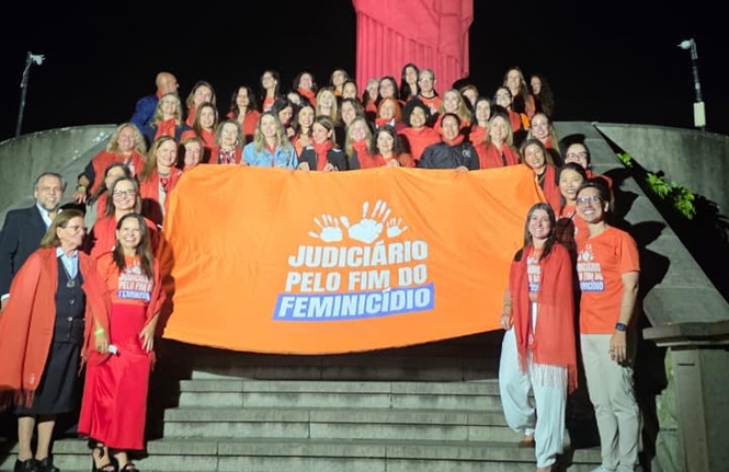 Foto horizontal: magistradas, servidoras e servidores posam reunidos em escadaria, à noite, segurando faixa da campanha "Judiciário pelo Fim do Feminicídio", diante de monumento iluminado na cor laranja em apoio à mobilização pelo enfrentamento da violência contra a mulher.