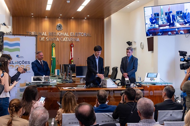 Foto horizontal: autoridades acompanham cerimônia no plenário do Tribunal Regional Eleitoral de Santa Catarina, durante demonstração de equipamentos e procedimentos preparatórios para as Eleições 2026, diante de convidados e profissionais da imprensa.
