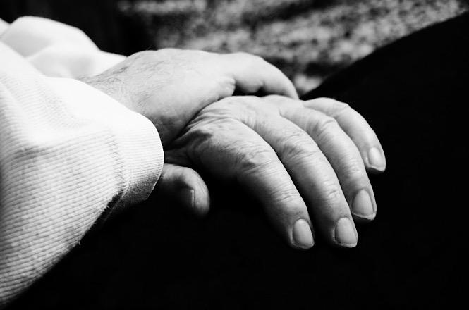 Foto horizontal em preto e branco: uma pessoa segura delicadamente a mão de outra, em gesto de acolhimento, cuidado e apoio, em imagem ilustrativa que representa empatia, solidariedade e conforto emocional.