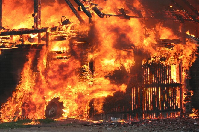 Foto horizontal: estrutura de madeira é consumida por um incêndio de grandes proporções, com intensas chamas e fumaça, em imagem ilustrativa que representa sinistros, destruição e atuação de emergência.