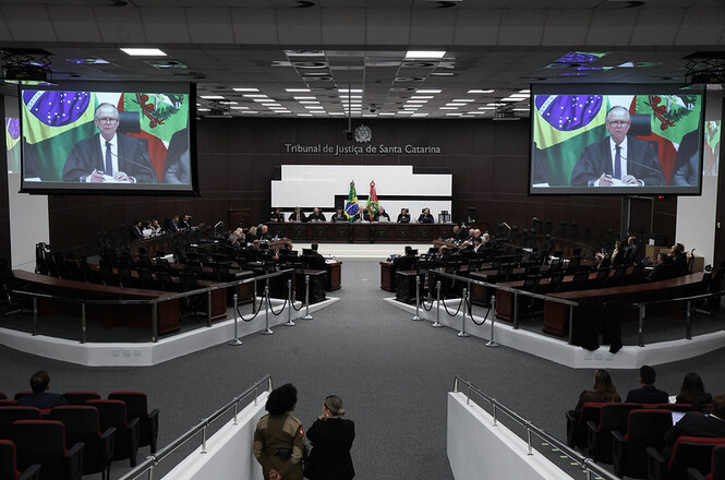 Foto horizontal: vista ampla do Pleno do Tribunal de Justiça de Santa Catarina durante sessão solene. Em dois telões, o desembargador Rubens Schulz aparece em pronunciamento transmitido ao público presente.