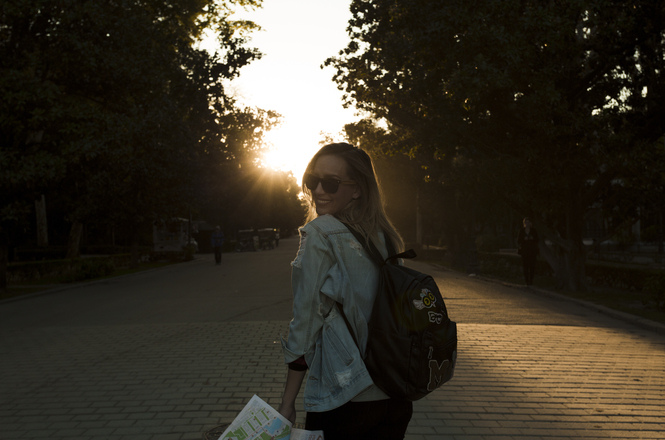 Foto horizontal: mulher caminha por via arborizada ao pôr do sol, carregando mochila e mapa, em imagem ilustrativa sobre deslocamento e mobilidade urbana.