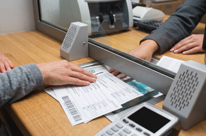 Foto horizontal: atendimento em guichê bancário, com entrega de boleto e dinheiro, em imagem ilustrativa sobre pagamentos e serviços financeiros.