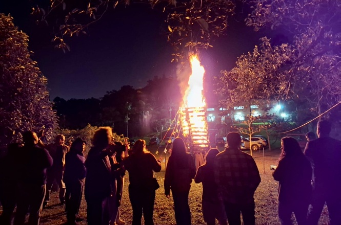 Foto horizontal: Pessoas acompanham fogueira acesa durante celebração ao ar livre em evento noturno.