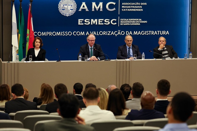 Foto horizontal: Magistrados participam de mesa de abertura durante evento realizado na Esmesc, com público no auditório.