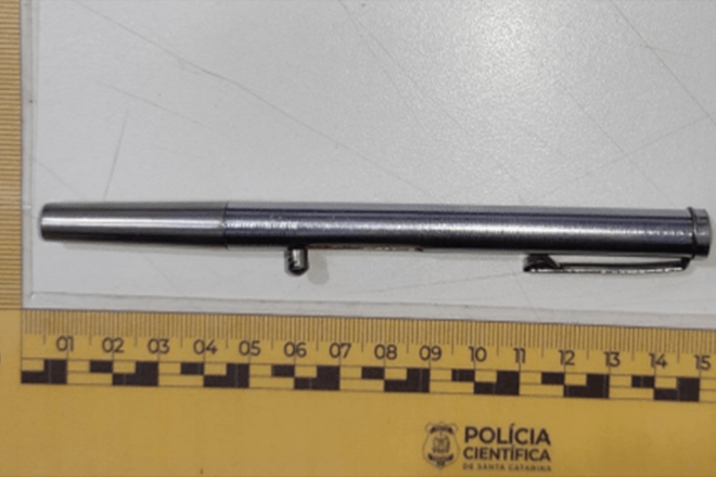 Foto horizontal: Caneta metálica apreendida e periciada pela Polícia Científica, posicionada sobre escala de medição.