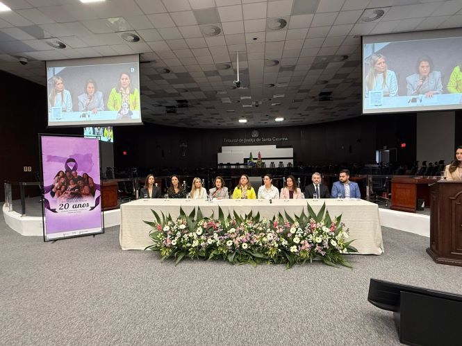 Fotografia panorâmica do auditório do Tribunal de Justiça de Santa Catarina durante o Seminário Lei Maria da Penha – 20 anos. No centro do palco, há uma mesa comprida ocupada por diversas pessoas, entre elas participantes e autoridades do evento. À frente da mesa, um grande arranjo de flores com folhagens e flores em tons de branco, rosa e lilás. À esquerda, há um painel vertical com a identidade visual dos 20 anos da Lei Maria da Penha. Duas telas grandes, suspensas nas laterais do auditório, exibem imagens em tempo real das participantes sentadas à mesa. Ao fundo, vê-se a identificação “Tribunal de Justiça de Santa Catarina” e as bandeiras institucionais. À direita, uma mulher está em pé junto ao púlpito, enquanto o restante dos participantes acompanha a atividade. O auditório é amplo, com iluminação no teto e cadeiras dispostas para o público.