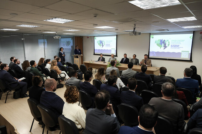 Foto horizontal: Vista ampla de auditório com participantes sentados diante de mesa de debatedores e projeções.