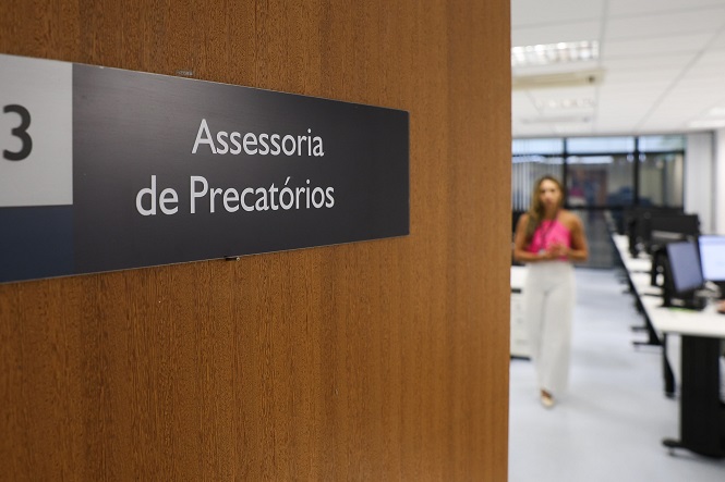 Foto horizontal: Placa identifica a Assessoria de Precatórios na entrada de setor, com ambiente de trabalho ao fundo.
