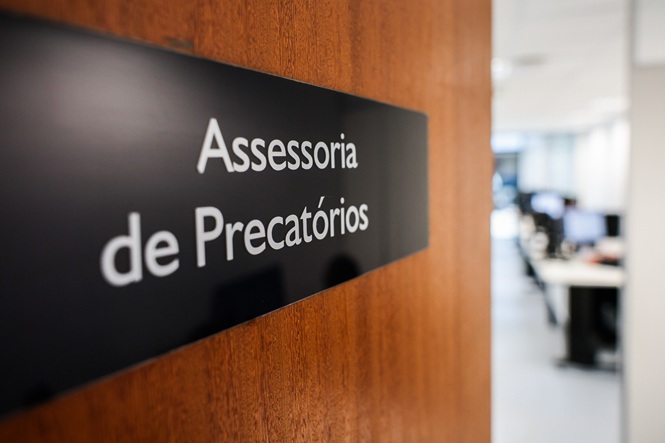 Foto horizontal: Placa identifica a Assessoria de Precatórios na entrada de setor, com ambiente de trabalho ao fundo.