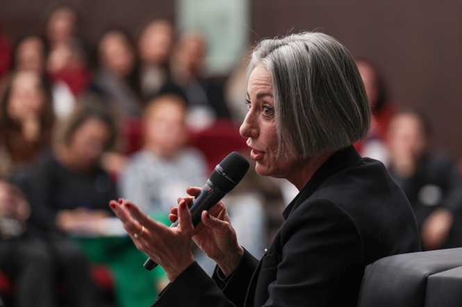Foto horizontal: Vera Iaconelli fala ao microfone durante evento, diante do público presente no auditório.