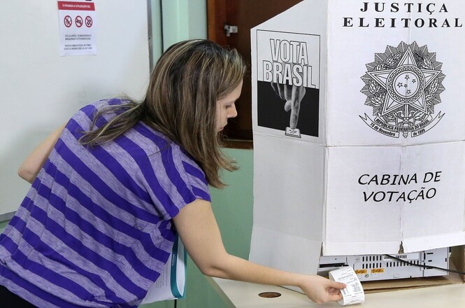 Foto horizontal: voluntária retira comprovante de votação em urna eletrônica da Justiça Eleitoral.