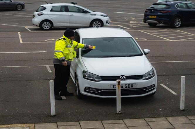 Foto horizontal: agente coloca aviso no para-brisa de veículo estacionado em área de estacionamento.