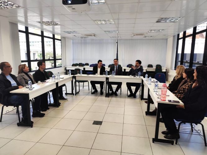 Fotografia de uma reunião realizada em uma sala ampla e iluminada, com participantes sentados ao redor de mesas dispostas em formato de “U”. Ao centro, três homens estão sentados, sendo que um deles fala enquanto os demais acompanham. À esquerda, há quatro participantes, e, à direita, outras quatro pessoas. Sobre as mesas estão garrafas de água, copos, blocos de anotações e outros materiais. Ao fundo, há janelas amplas com persianas, cadeiras e uma bandeira.