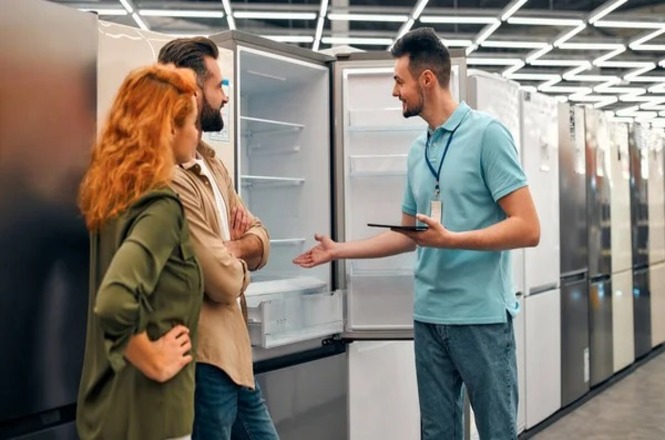 Foto horizontal: vendedor apresenta eletrodoméstico a clientes em loja, diante de refrigeradores