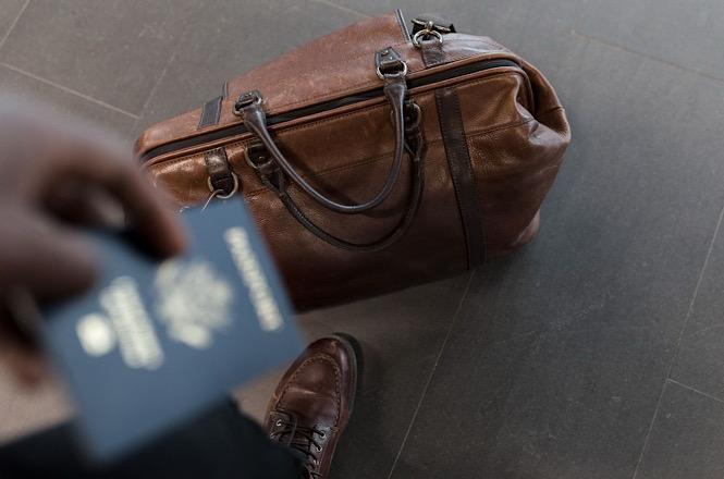 Foto horizontal: mala de viagem marrom ao lado de pessoa que segura um passaporte.