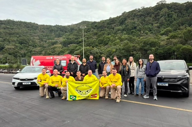 Foto horizontal: grupo reunido em pista de treinamento, com veículos e equipe de pilotagem.