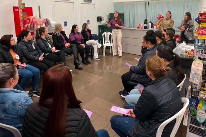 Foto horizontal: mulheres participam de roda de conversa em estabelecimento comercial, com palestrante ao centro.
