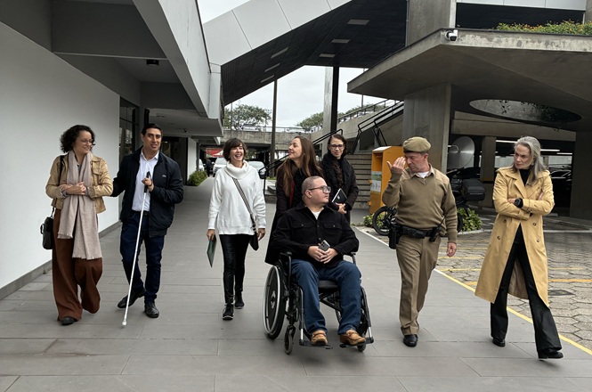 Foto horizontal: grupo caminha em área externa, com pessoas com deficiência visual e cadeirante.