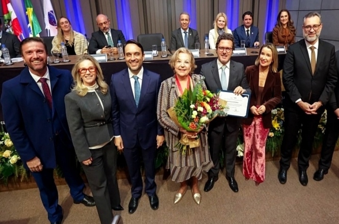 Foto horizontal: autoridades posam com homenageada que segura flores durante solenidade institucional.