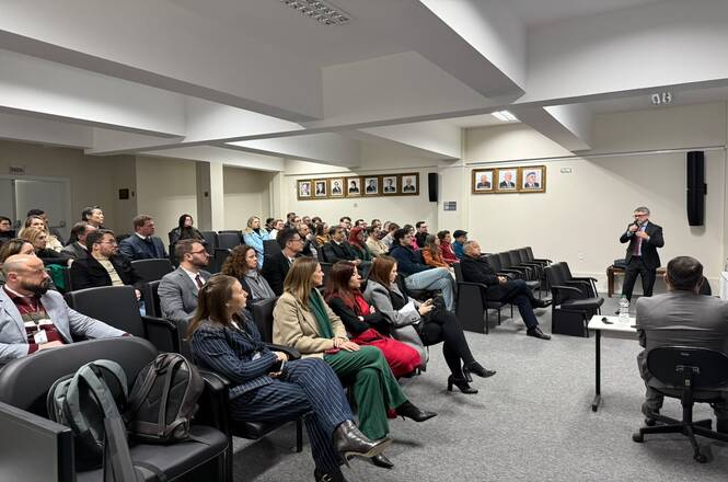 Foto horizontal: público acompanha palestra em auditório, com palestrante falando à frente dos participantes.