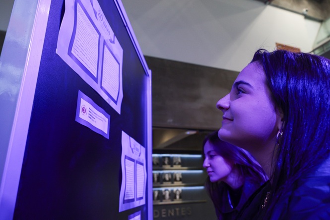 Imagem horizontal mostra mulher observando documentos expostos em painel, sob iluminação azul.
