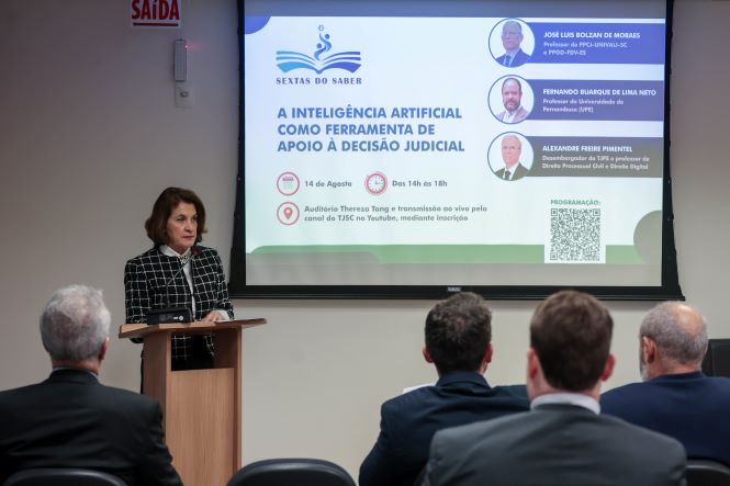 Fotografia de uma palestra realizada em auditório. Em primeiro plano, a desembargadora Vera Copetti está ao púlpito, falando ao público. Ela veste blazer preto com padrão xadrez branco. À frente, há pessoas sentadas de costas, acompanhando a apresentação. Ao fundo, um telão exibe a programação do “Sextas do Saber”, com o tema “A Inteligência Artificial como ferramenta de apoio à decisão judicial”, além dos nomes dos três palestrantes: José Luiz Bolzan de Moraes, Fernando Albuquerque de Lima Neto e Alexandre Freire Pimentel.