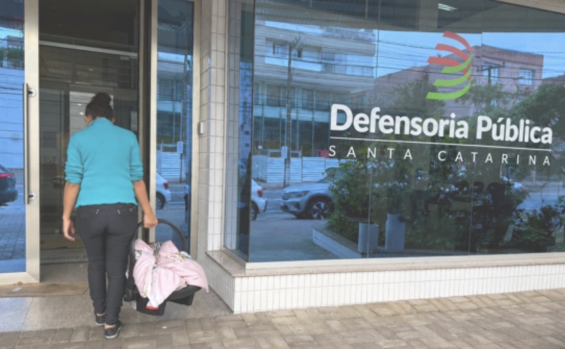 Imagem horizontal mostra mulher entrando na Defensoria Pública de SC, carregando bolsa e bebê-conforto.