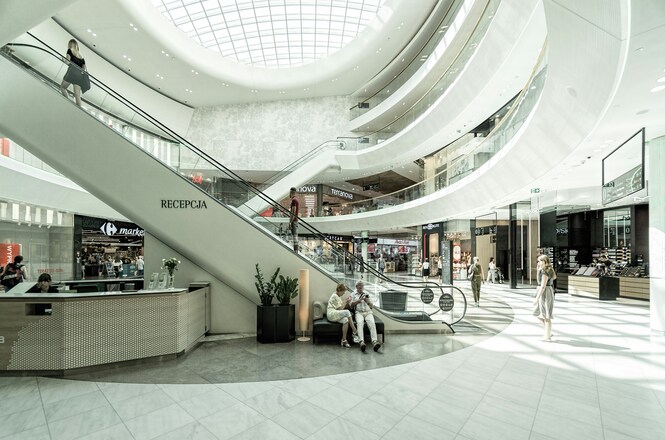 Imagem horizontal mostra interior amplo de shopping center, com escadas rolantes, lojas e pessoas circulando.