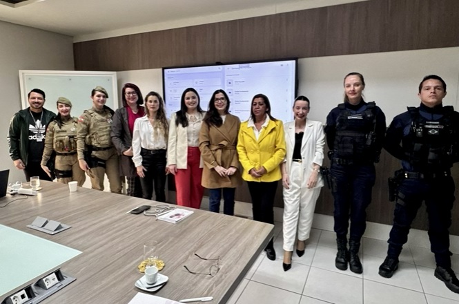 Imagem horizontal mostra autoridades, policiais militares e agentes de segurança reunidos em sala institucional.
