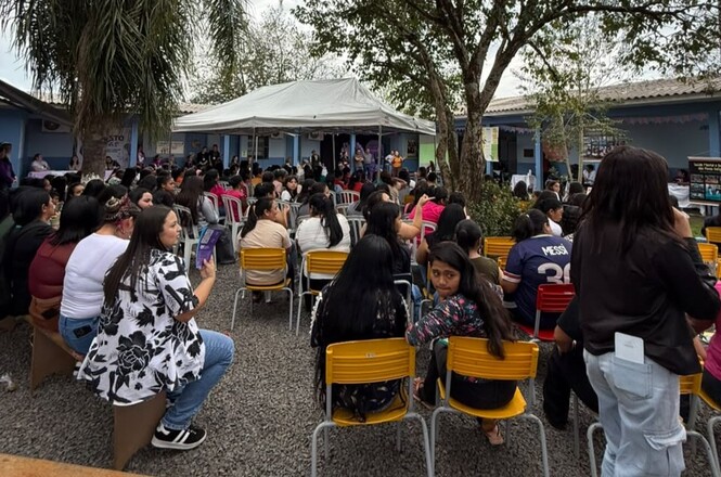 Imagem horizontal mostra público reunido em área externa, sentado em cadeiras durante evento comunitário.