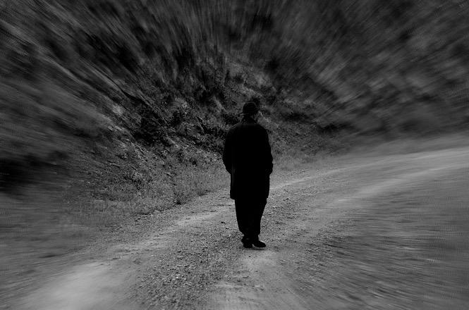 Imagem horizontal em preto e branco mostra pessoa caminhando sozinha por estrada de terra, em cenário desfocado.