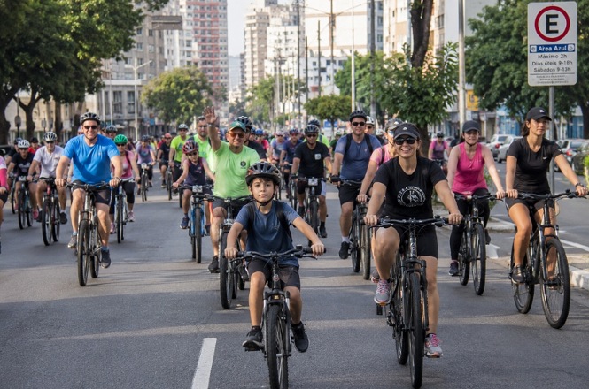 Imagem horizontal mostra ciclistas de diferentes idades pedalando em grupo por avenida urbana.