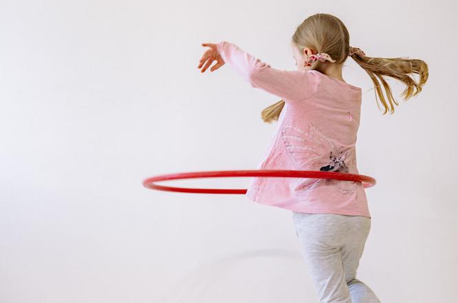 Imagem horizontal mostra criança brincando com bambolê vermelho, em movimento diante de fundo branco.