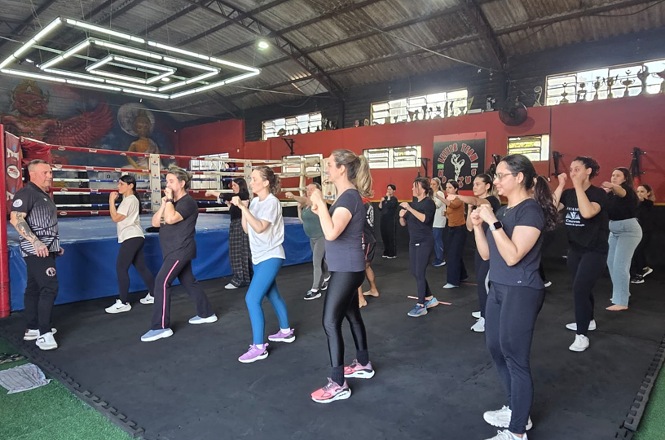 Imagem horizontal mostra grupo de mulheres praticando movimentos de defesa pessoal em academia com ringue de boxe.