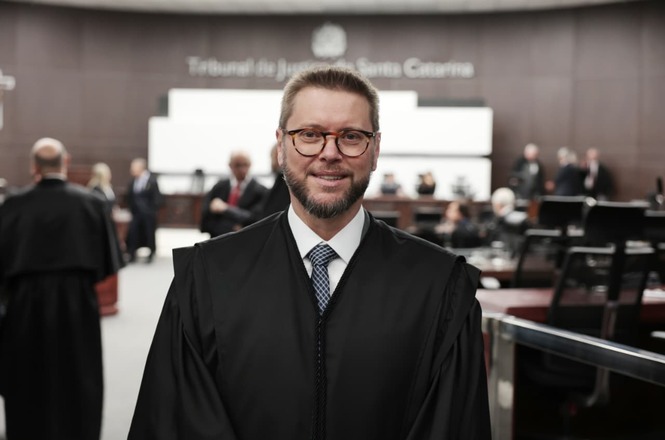 Imagem horizontal mostra o magistrado Giancarlo Bremer Nones de toga, posando no plenário do Tribunal de Justiça de Santa Catarina.