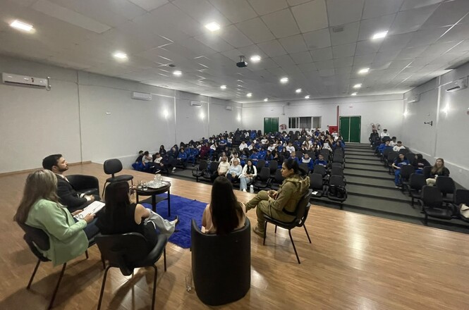 Imagem horizontal mostra palestrantes em roda de conversa diante de estudantes reunidos em auditório.