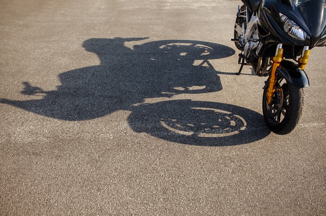 Imagem horizontal mostra motocicleta estacionada no asfalto, com sua sombra projetada pela luz do sol.
