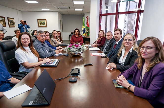 Imagem horizontal mostra autoridades reunidas em torno de mesa, durante encontro institucional em sala.