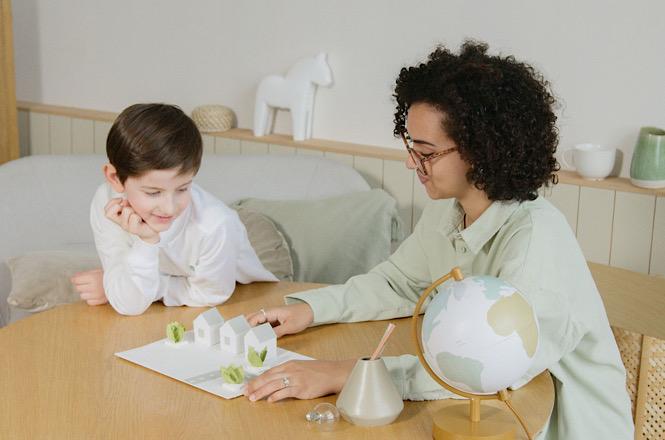 Imagem horizontal mostra mulher e criança realizando atividade educativa com maquete e globo terrestre.